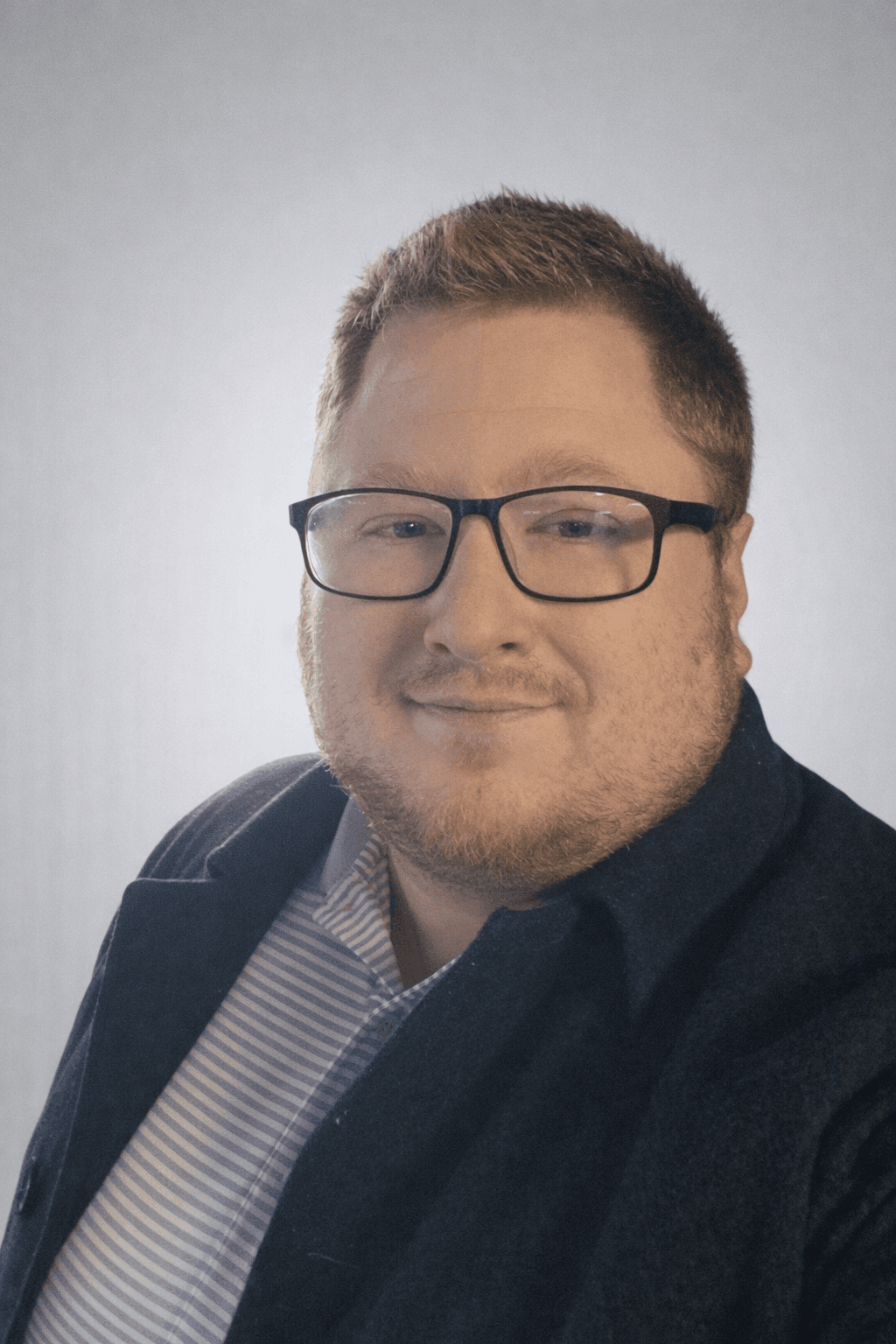 Smiling man with short reddish hair, glasses, and beard wearing a blazer and striped shirt.